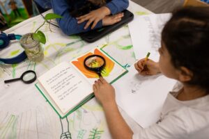 Innovations Academy student holds a pencil in one hand and the other has a magnifying glass held over an illustration in a book.