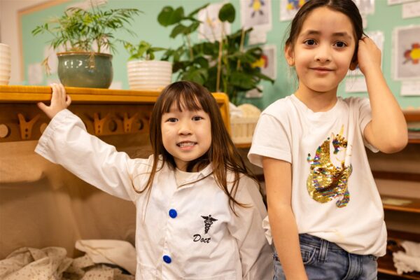 Two Innovations Academy students in a classroom with plants and wooden furniture look at the camera, one in a dress-up doctor jacket.