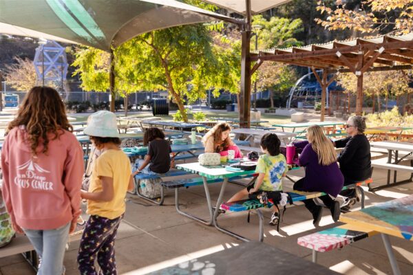 Adults and students at Innovations Academy (IA) sit and stand around colorfully decorated long picnic tables outside under soaring sunshades.