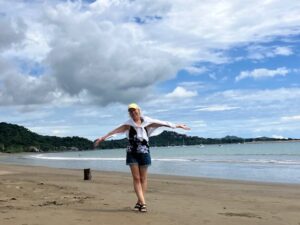 Innovations Academy founder Christine Kuglen stands with outstretched arms on a curving beach of sand with hills forested with trees in the background as a few shallow waves crash to shore.