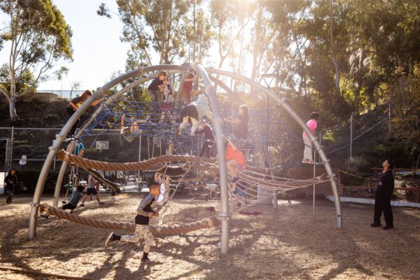 Innovations Academy students play on a bridge and net structure and run around outside with shady trees overhead.
