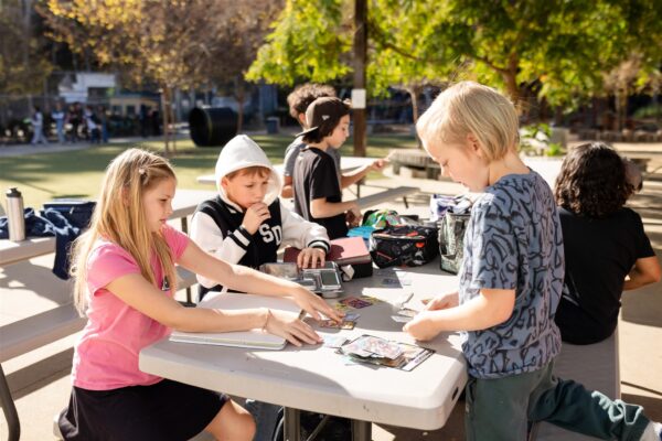 Innovations Academy students sit at tables with papers on them outside in a tree-filled, sun-dappled area.