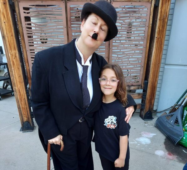 Innovations Academy (IA) Performing Arts teacher Natasha Starbuck-Smith poses in a suit and bowler hat costume complete with a mustache and cane and side hugs an IA student.