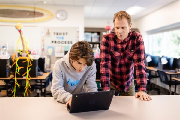 A student hovers over a laptop while a teacher stands next to them looking over their shoulder at Innovations Academy.
