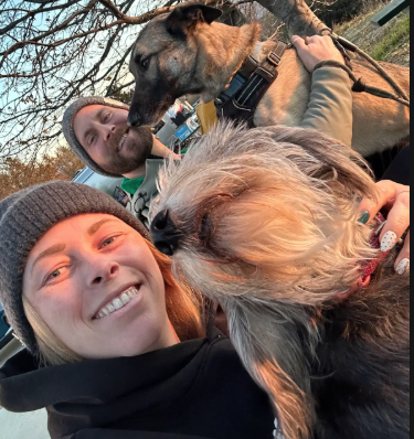 Innovations Academy (IA) Language Arts teacher Devon smiles outside under a tree holding a smaller shaggy dog while a person behind her has a bigger dog with their front legs on their lap.
