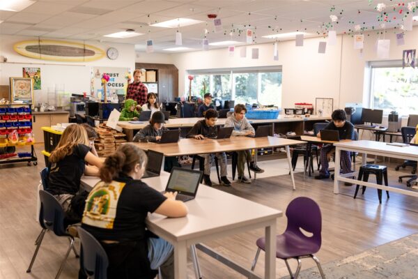 Middle school students in an Innovations Academy airy classroom sit at tables working on laptops. 