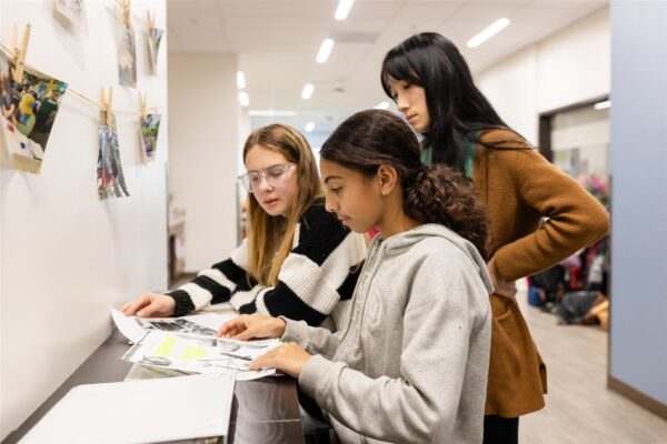 nnovations Academy (IA) students sit at a long, narrow high top table looking through illustrated papers.