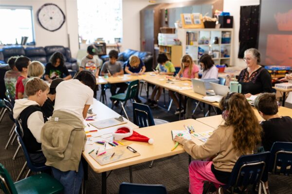 Innovations Academy (IA) students and an adult sit at long tables formed into a rectangle with colorful pens and pencils laid out along with paper and clipboards.