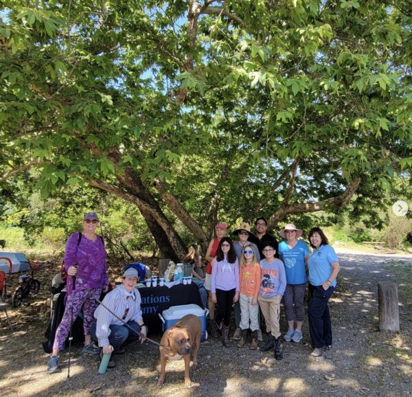 Innovations Academy (IA) students and adults, along with one large brown dog, stand flanking an Innovations Academy table under a large tree