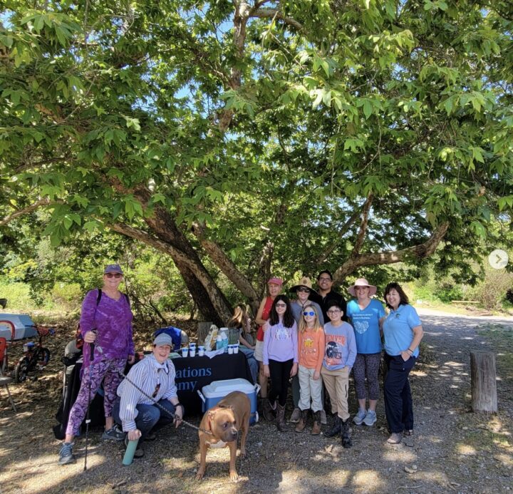 Innovations Academy (IA) students and adults, along with one large brown dog, stand flanking an Innovations Academy table under a large tree