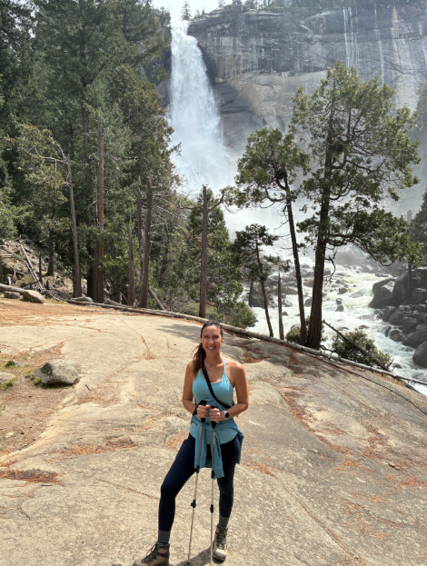 Innovations Academy (IA) teacher Kristy Fitts stands on a large rock boulder in the foreground with hiking poles while the background is taken up by a multi-story waterfall breaking onto a rocky channel below with trees around.