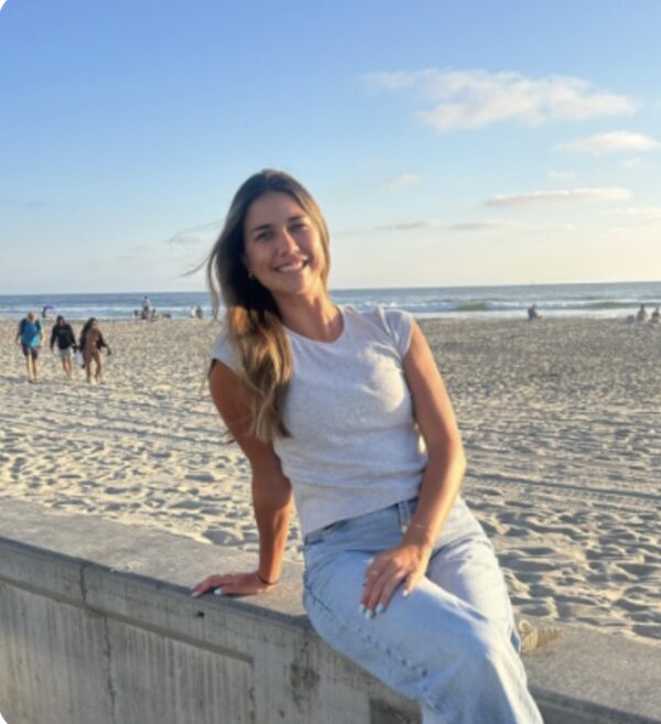 Innovations Academy (IA) teacher Riley Gray sits on a concrete wall with a sandy flat beach and ocean behind her.