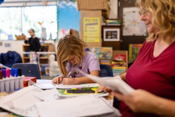 Innovations Academy (IA) student and adult sit at a classroom table with papers all over it and some markers. The student is bent over their paper coloring with the marker.