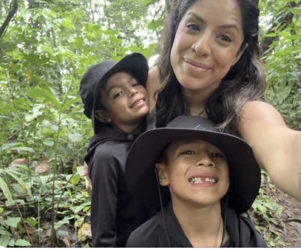Innovations Academy (IA) teacher Jackie Pina smiles with two small children all in black clothing in a forested setting.