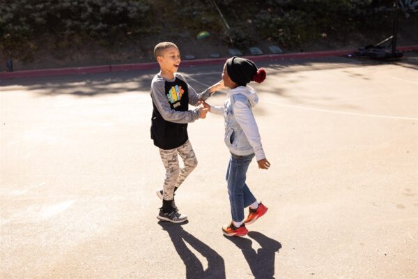 Innovations Academy (IA) students greet each other with huge smiles mid-step outside in the sunshine.