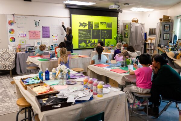Innovations Academy (IA) students sit in a classroom with dropsheets over the tables and look at a teacher who is writing on the whiteboard to the left of a neon green screen with black squares and shapes.
